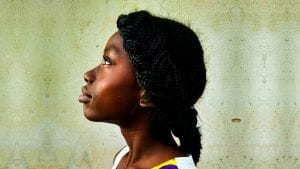 Am I Cursed? Zambian woman looking up with white wall behind.